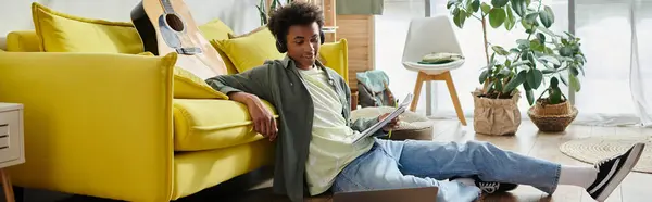 A young African American man sits on a yellow couch, focused on his laptop screen. — стоковое фото