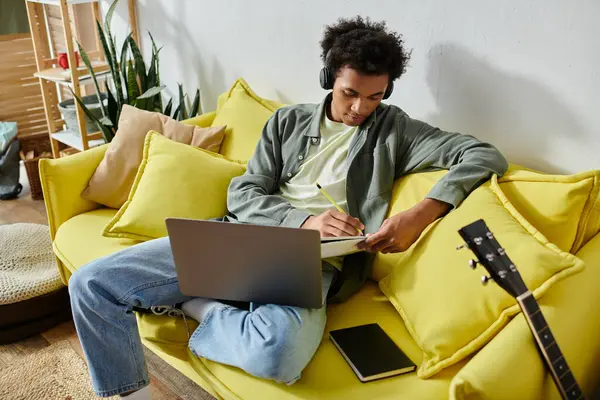 A young African American man sits on a yellow couch with headphones and a laptop. — Stock Photo