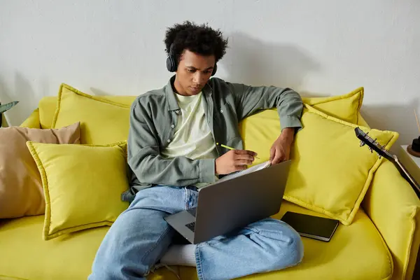 Young African American man studying with headphones and laptop on yellow couch. — Stockfoto