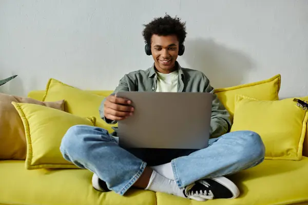 Young African American man engrossed in online study, seated on yellow couch with laptop. - foto de stock