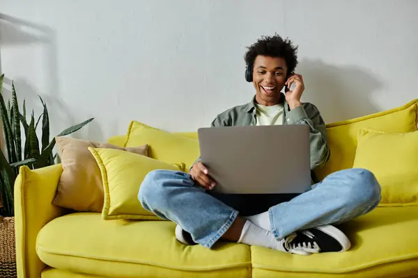Young African American man studying online on a yellow couch with a laptop. — Foto stock