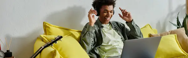 Young man, African American, sitting on yellow couch, using laptop for online study. — Stockfoto