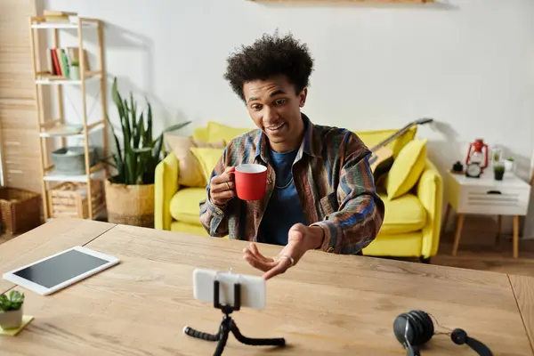 A young African American male blogger holding a cup of coffee while talking on a phone camera at a table in front of a yellow couch. — Photo de stock