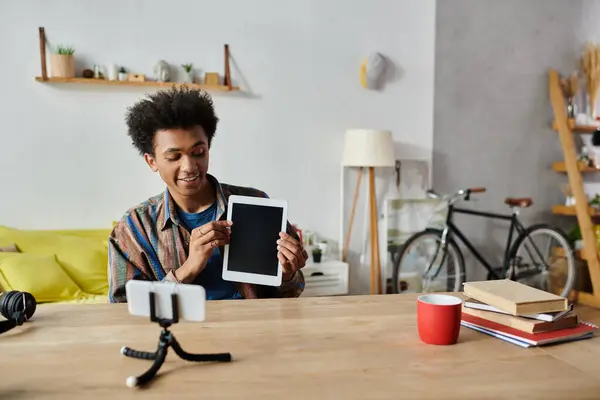 A young African American male blogger is sitting at a table with a tablet in front of him, discussing into a phone camera. — стокове фото