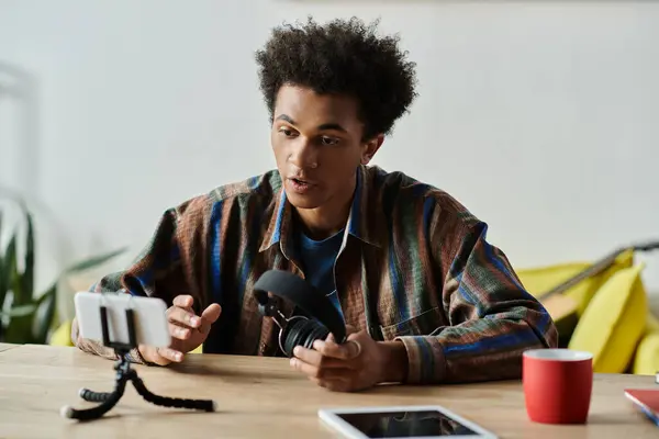 A young African American man sits at a table, engaging with a phone and tripod while talking. — стоковое фото