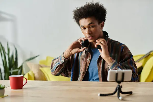 A young African American blogger sits at a table, savoring a cup of coffee while speaking into his phone camera. — Stock Photo