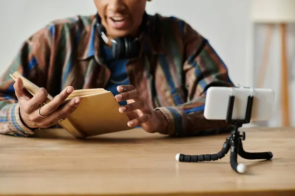 A young African American man reads a book while conversing on his phone camera at a table. — Stock Photo