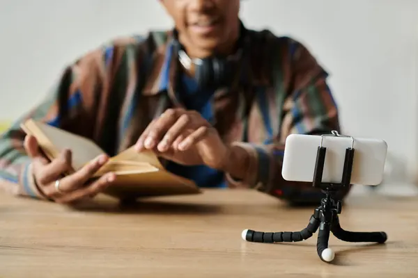 African American man reads book while vlogging on phone tripod. — стокове фото