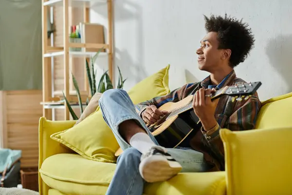 A young African American man plays an acoustic guitar while seated on a yellow couch. — Stock Photo
