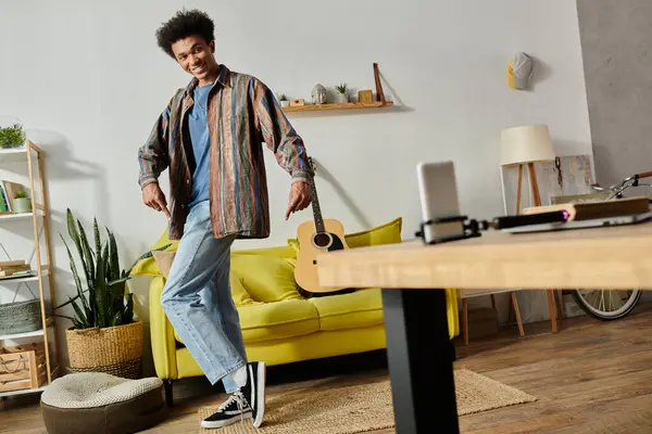 Young African American male blogger playing guitar, talking on phone camera in front of yellow couch. — Stock Photo
