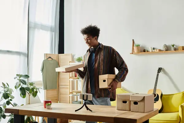 A young African American male blogger is holding a box in front of a table while talking on a phone camera. — Stock Photo