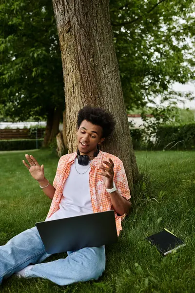 A young African American man sits on the grass, engrossed in working on his laptop. - foto de stock