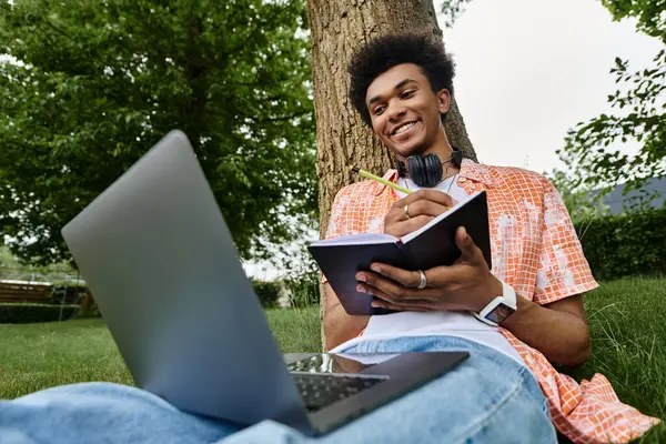 Young man, African American, sitting under tree, using laptop and headphones in park. — Stock Photo