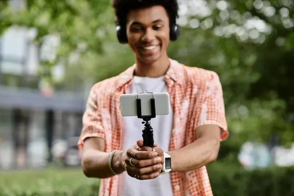 A young African American man holds a selfie stick in a park. — Stock Photo