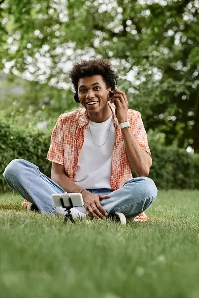 Young man, African American, sitting on grass, listening to music on phone. — Stock Photo