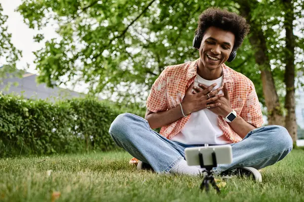 Young African American man sitting on grass, engrossed in cell phone. — Fotografia de Stock