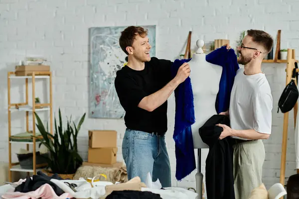 Dos hombres, una pareja gay, están uno al lado del otro en un taller de diseño, colaborando en atuendos de moda. - foto de stock
