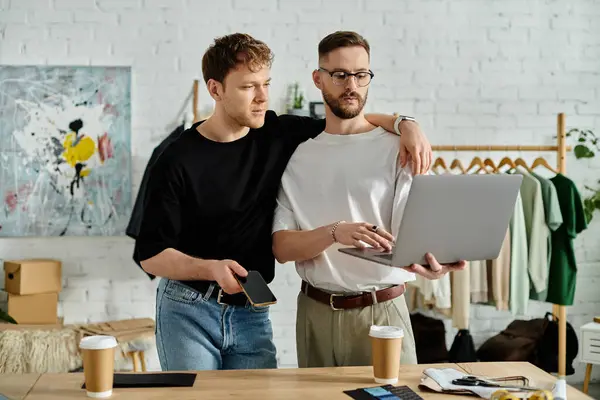 Una coppia gay sta fianco a fianco di fronte a un computer portatile, lavorando appassionatamente insieme su abiti alla moda in un laboratorio di design. — Foto stock