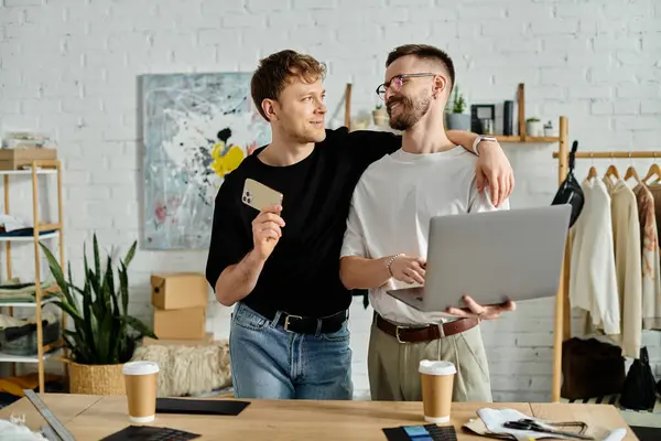 Dos hombres, una pareja gay, están en un taller de diseño, enfocados en la pantalla de un portátil mientras discuten la creación de atuendos de moda. — Stock Photo