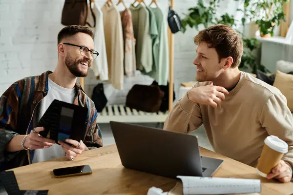 Two men, a gay couple, work together at a table with a laptop, designing trendy attire in a designer workshop. — Stock Photo