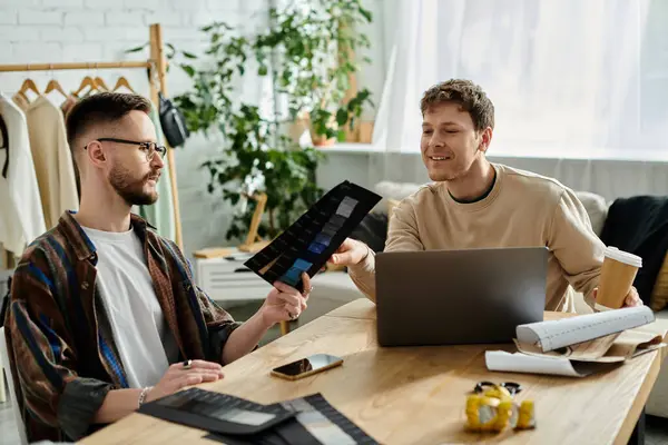 Two men, a gay couple, sit at a table with a laptop, collaborating on trendy attire they design together in their workshop. — Stock Photo