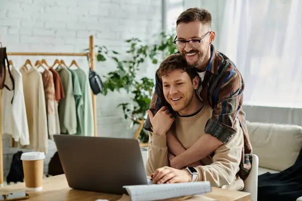 Um homem abraça seu parceiro como eles colaboram na concepção de trajes na moda em meio a um laptop. — Fotografia de Stock