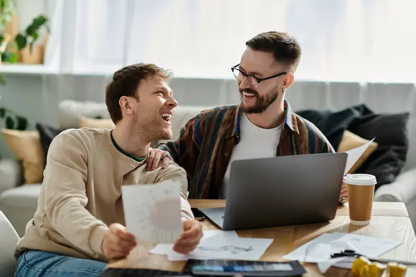 Zwei Männer, Teil eines schwulen Paares, sitzen an einem Tisch, vertieft in ihren Laptop-Bildschirm, während sie gemeinsam trendige Kleidung entwerfen.. — Stockfoto