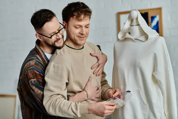 A gay couple stands side by side in a designer workshop, working together on trendy attire. — Stock Photo