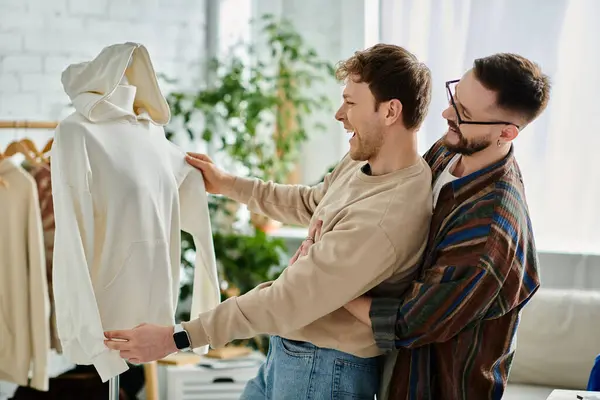Two men, a gay couple, stand side by side, admiring a completed trendy attire in a designer workshop. — Photo de stock