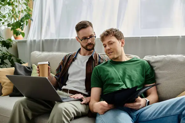 Dos hombres enamorados se sientan en un sofá, enfocados en un portátil, diseñando atuendos de moda en su taller. - foto de stock