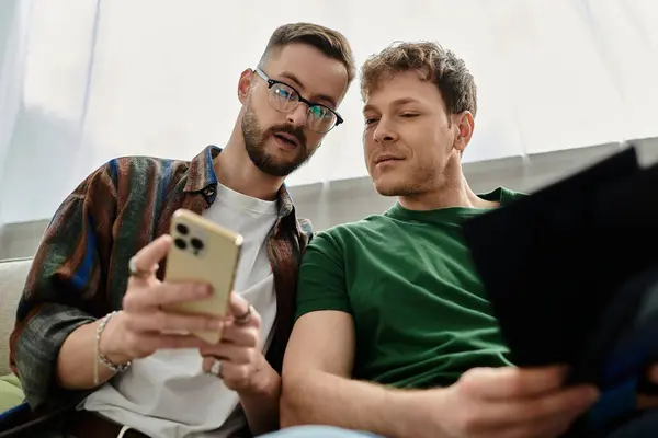 Two men in love sit on a couch, focused on a cell phone screen. — Stock Photo