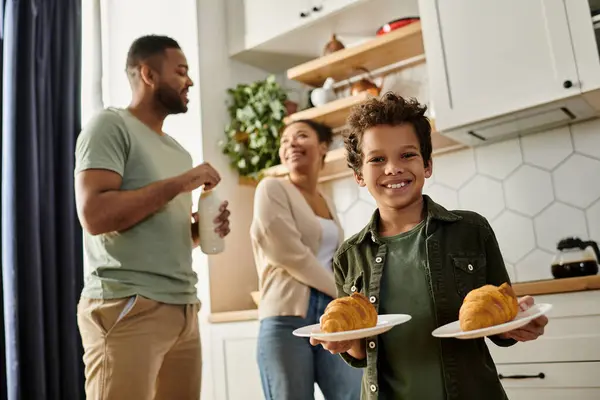 Un niño sostiene cuidadosamente dos platos de comida con sus padres en el fondo. - foto de stock