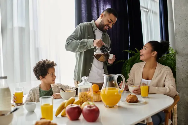 Un hombre vierte jugo de naranja para una amorosa familia afroamericana en la mesa del desayuno. - foto de stock