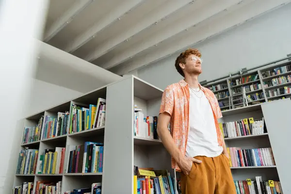 Junger Mann steht vor Bücherregal voller Bücher in Bibliothek. — Stockfoto