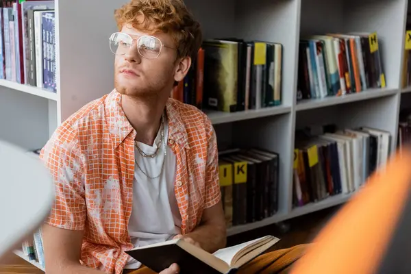 A young man engrossed in reading a book while seated in front of a bookshelf. — Stock Photo