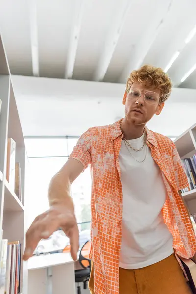 A young, handsome male student standing in front of a bookshelf in a library. — Stock Photo