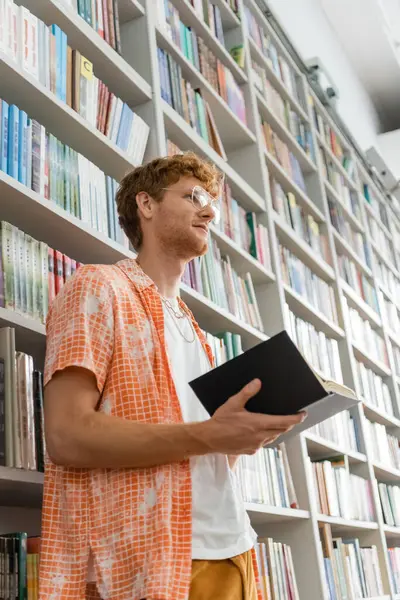 A young man stands before a bookshelf, holding a book. — Stock Photo