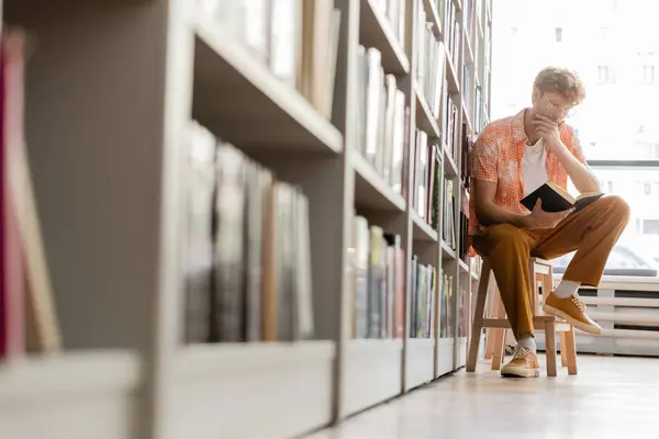 A young man sits before a bookshelf, deep in study. — Stock Photo