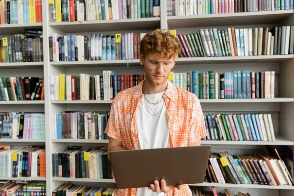 Young man with laptop at a bookshelf in library. — Stock Photo