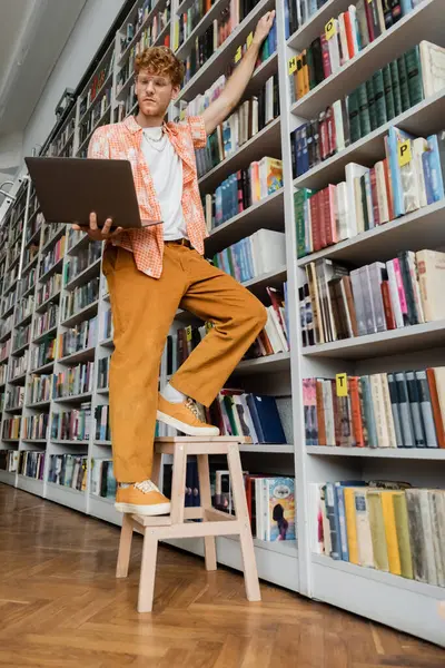 Юнак стоїть на табуреті перед книжковою полицею. — Stock Photo