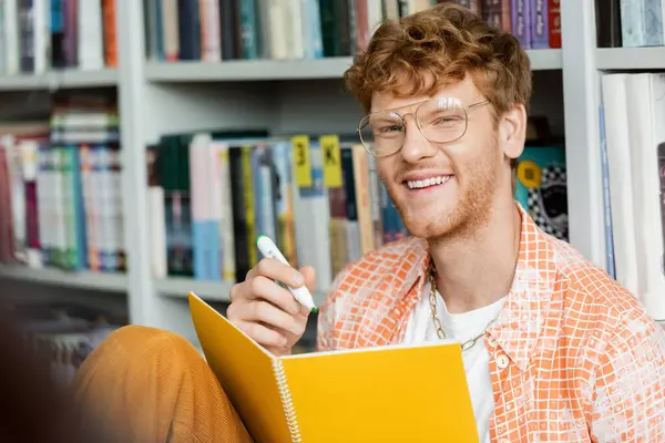 A young man seated before a bookshelf with a pen and notebook. — Stock Photo