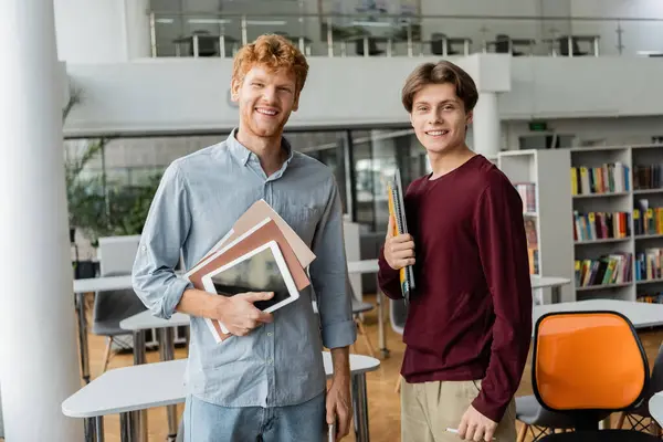 Two young male students stand side by side, each holding a book in a library. — Stock Photo