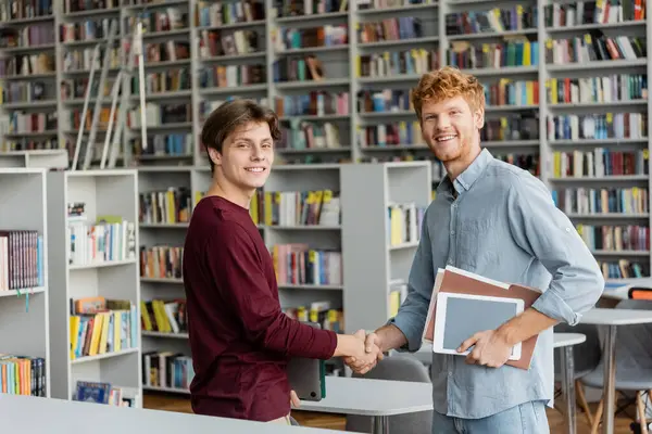Two young men shake hands in a quiet library, surrounded by books and studying materials. — Stock Photo