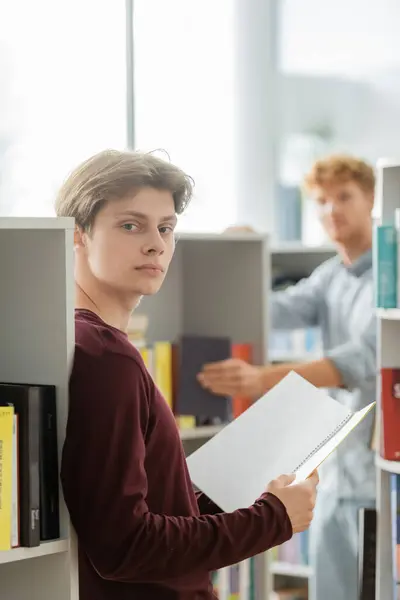 Man holding a book in front of bookshelf, with his friend on backdrop. — Stock Photo