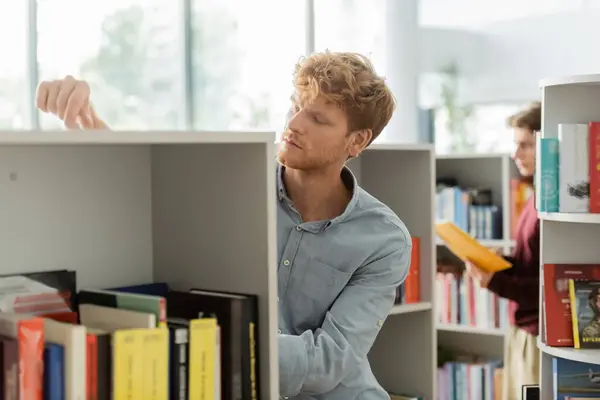 A man casually leaning against a bookshelf in a library, with his friend on backdrop. — Stock Photo