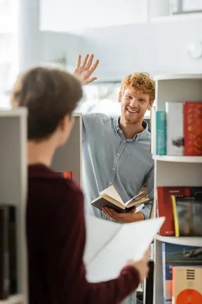 A man stands in front of a bookshelf and his friend, holding a book. — Stock Photo