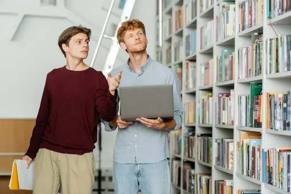 Two men engrossed in laptop research amidst stacks of books in a library. — Stock Photo