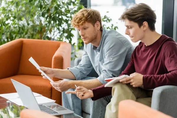 Two young men sitting on a couch, examining papers with focused expressions. — Stock Photo