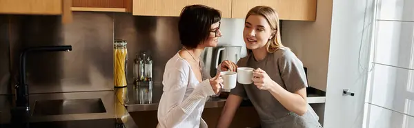 A young lesbian couple chat over coffee cups, standing together in a cozy hotel kitchen. — Stock Photo