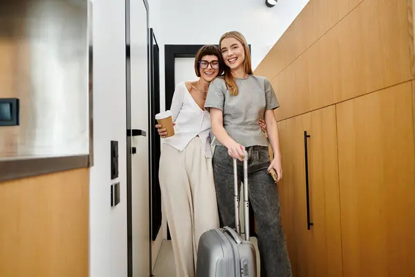 Um jovem casal lésbico está em um quarto de hotel, pronto para embarcar em uma viagem, segurando xícaras de café e bagagem.. — Fotografia de Stock